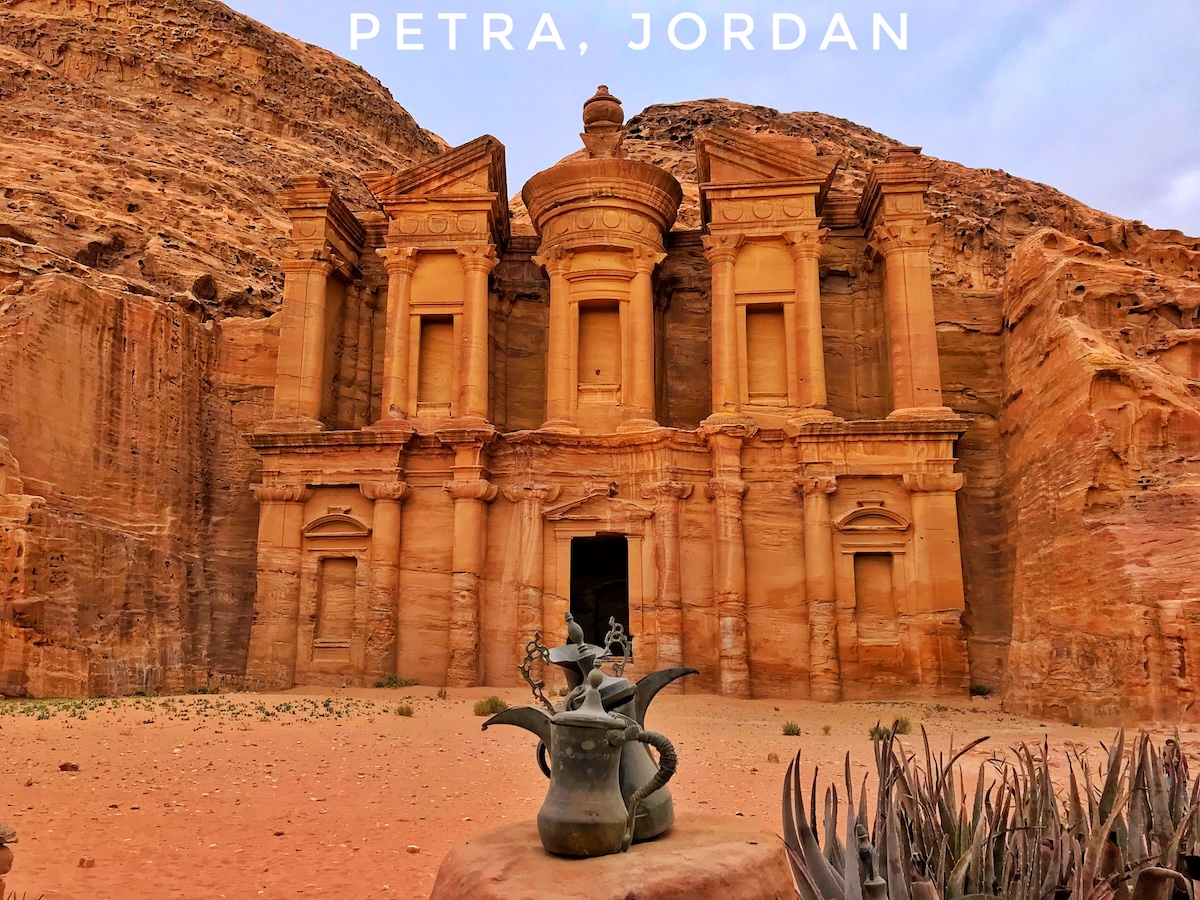 The Monastery (Al-Deir) in Petra, Jordan, a massive ancient building carved into sandstone cliffs, with a traditional teapot in the foreground, set against a backdrop of rugged desert landscape.