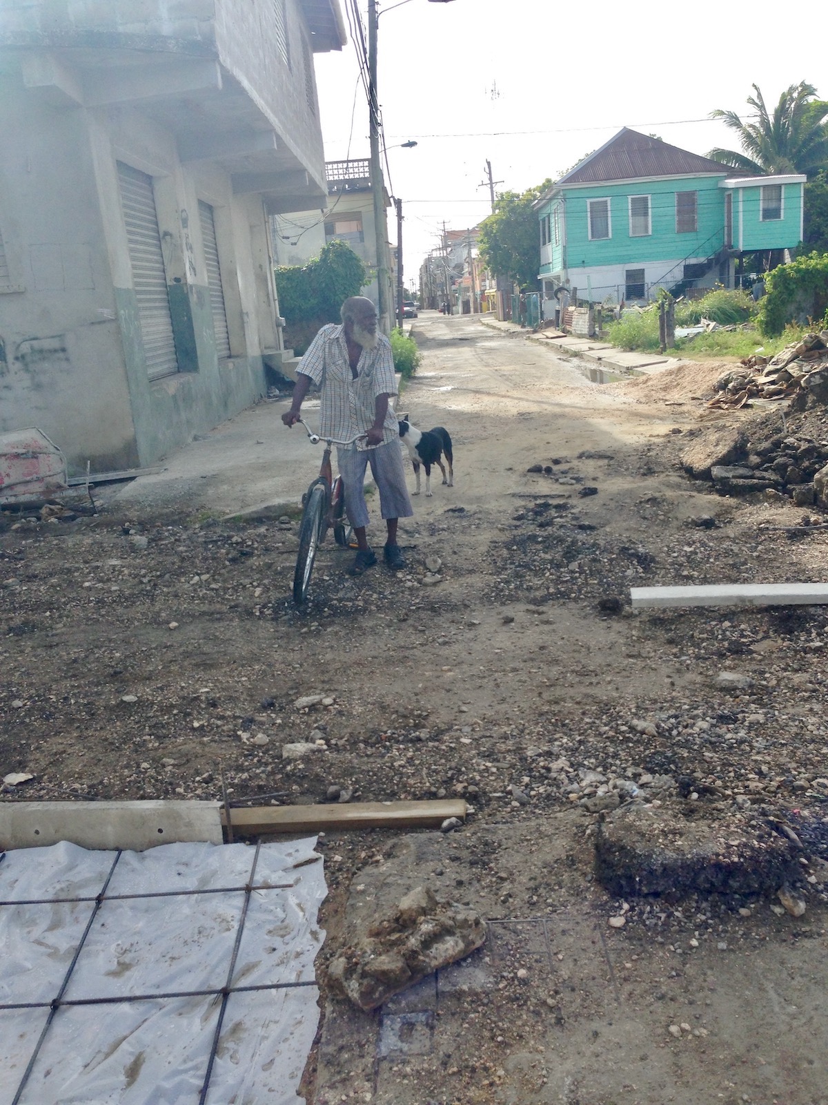 An old man with a long white beard walks with his dog and a bike in a gritty neighbourhood. 