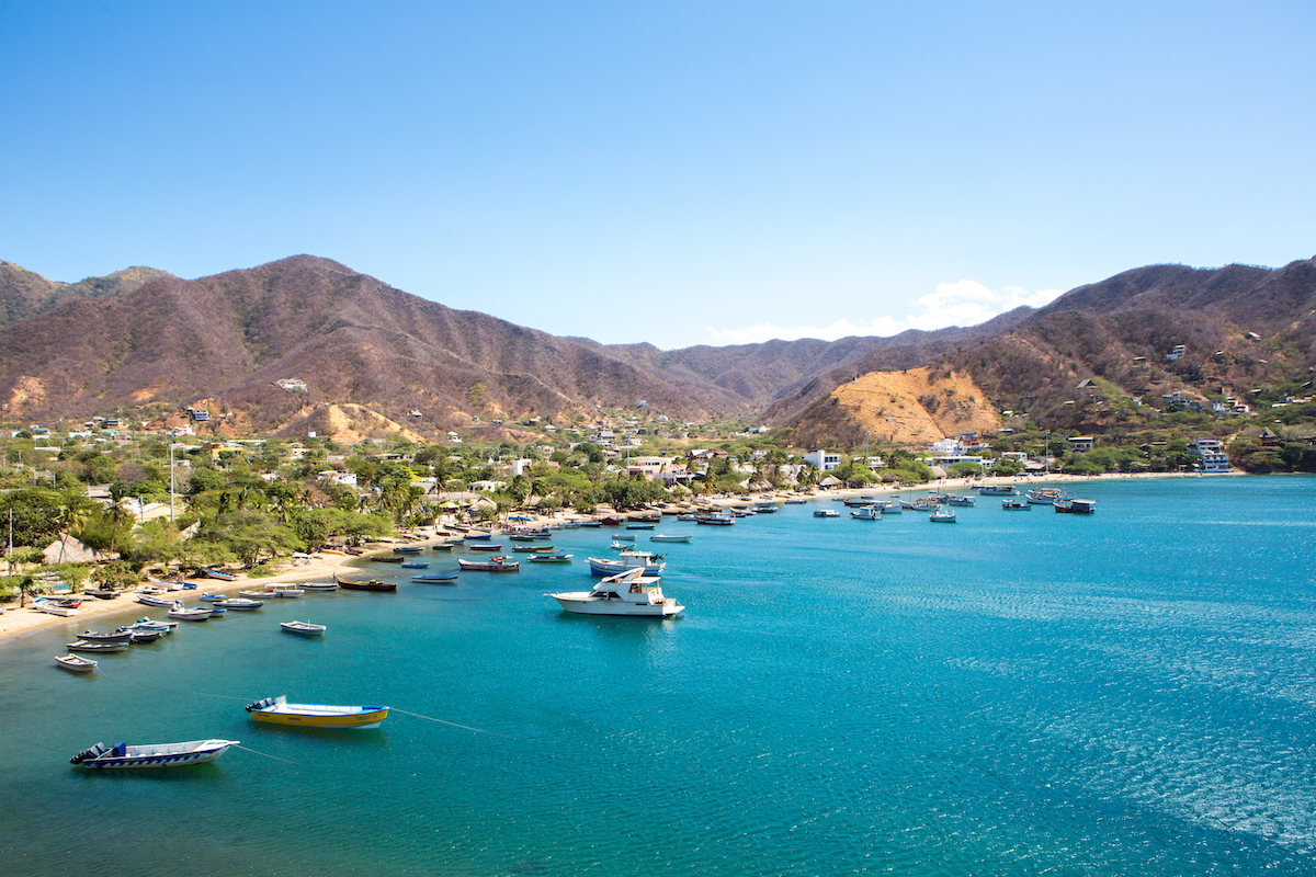 Boats sit on top of a blue clear sea overlooking brown hills in Taganga Bay, Colombia.